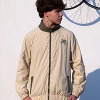 Person wearing a beige jacket with a logo, standing against a white wall with a bicycle.