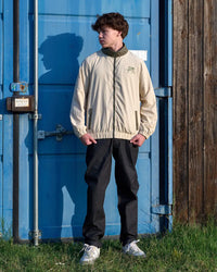 Person wearing a beige jacket with a logo standing in front of a blue container.