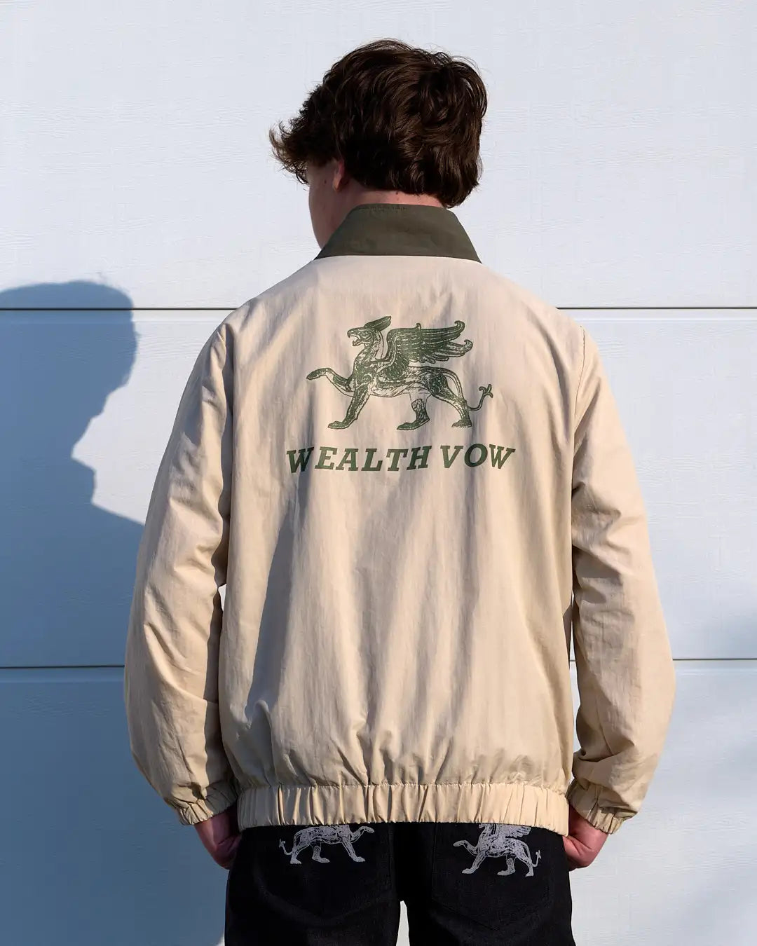 Person wearing a beige jacket with 'WEALTH VOW' logo on the back against a light blue wall.