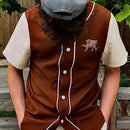 Person wearing a brown button up shirt with a logo, white shirt, and gray cap outdoors.